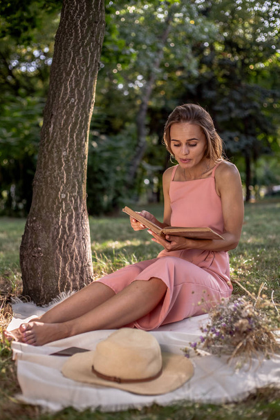 Bella ragazza che riposa sull'erba vicino all'albero. In un cappello con un libro di fiori secchi e uno smartphone
. - Foto, immagini