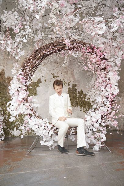 charming handsome Caucasian young man in a white business suit, jacket and trousers, posing against a pink Sakura. - Fotoğraf, Görsel