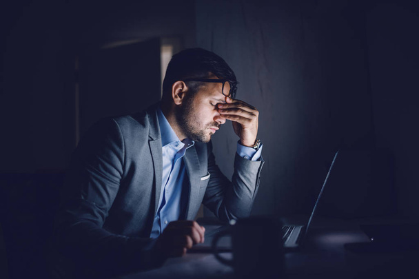Tired hardworking handsome Caucasian businessman sitting in office late at night and rubbing eyes. In front of his is laptop. Overworking concept. - Фото, зображення