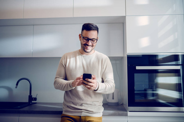 Atrakcyjny kaukaski uśmiechnięty człowiek ubrany casual opierając się na licznik kuchni i za pomocą inteligentnego telefonu, aby wysłać wiadomość. Wnętrze domu. - Zdjęcie, obraz