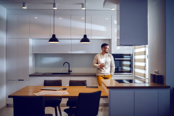 Yakışıklı Kafkas adam tam uzunlukta rahat mutfak tezgahına eğilerek giyinmiş, kahve içme ve yalak pencere görünümlü. Modern mutfak iç. - Fotoğraf, Görsel