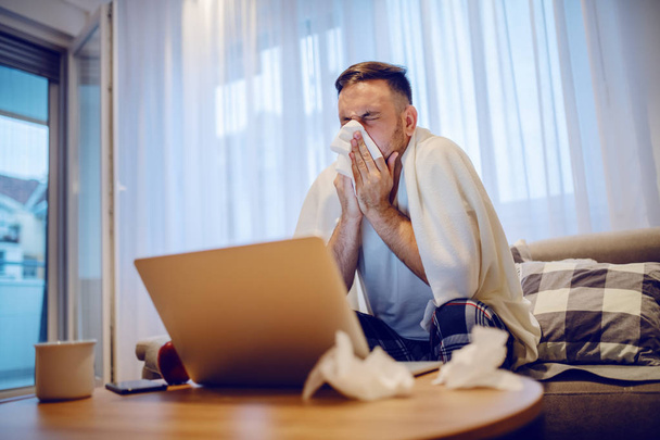Ill caucasian employee in pajamas sitting on the sofa in living room covered with blanket and blowing nose. On table are used handkerchiefs, laptop and mug with tea. - Photo, Image