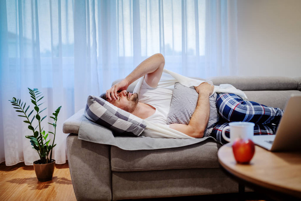 Very sick caucasian man in pajamas and covered with blanket lying on sofa in living room, holding pillow and having stomachache. - Foto, Imagen