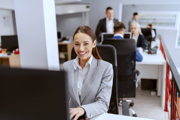 Beautiful positive Caucasian businesswoman in formal wear sitting in office and using computer. Hands on keyboard. In background colleagues working. - Фото, зображення