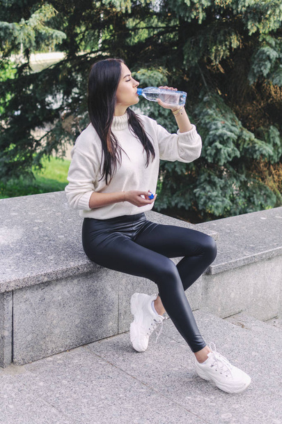 красива брюнетка дівчина п'є воду з пластикової пляшки, сидячи на мармуровому парапеті
 - Фото, зображення