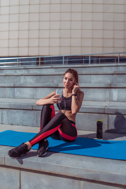 A beautiful woman athlete sits on a yoga mat, holds a phone, listens to music on headphones, a shaker with water and protein, rest after a workout, lifestyle workout fitness. Bright sunny day. - Фото, изображение