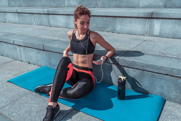 Beautiful girl athlete sits on yoga mat, holds phone, listens music headphones, watches video, listens podcast lecture chat message voice, rest after workout, lifestyle fitness. Bright sunny day. - Fotografie, Obrázek