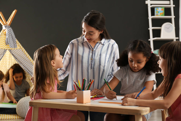 Cute little children with nursery teacher drawing at table in kindergarten. Indoor activity - 写真・画像