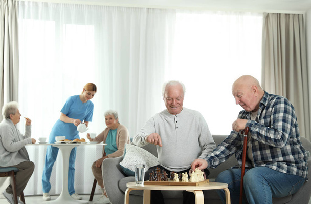 Starsi mężczyźni grają w szachy, podczas gdy pielęgniarka serwuje śniadania dla kobiet w domu emerytalnym. Pomaganie starszym ludziom - Zdjęcie, obraz