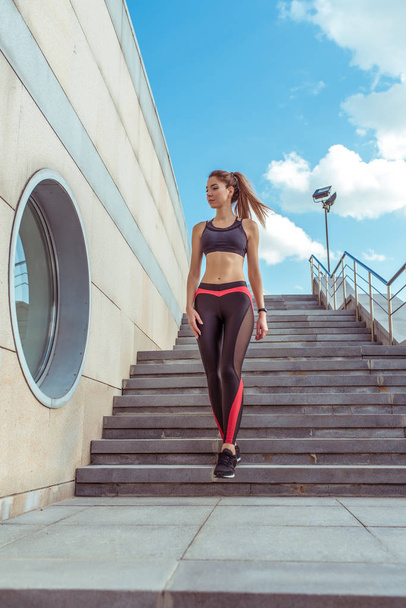 A beautiful girl, top sports leggings, walks stairs summer city a jog in morning, active lifestyle of young people. Fitness walk, workout, slim tanned figure. Clouds building staircase background. - Фото, зображення