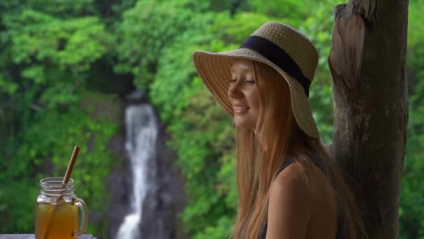 Slowmotion shot of a young woman in a beautiful cafe with an epic view on a canyon and waterfall drinks tea with ice. Travell to Bali concept - Πλάνα, βίντεο