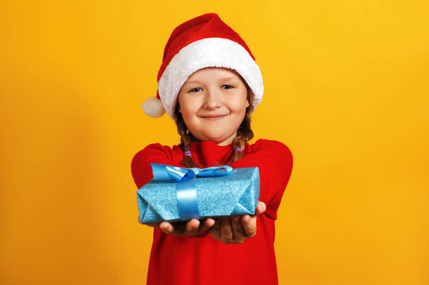 Happy little girl with christmas present. A child in a Santa hat gives a present - Фото, изображение