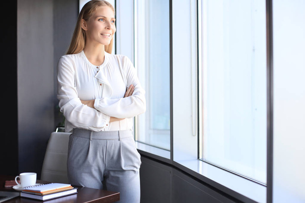 Attractive business woman looking at camera and smiling while standing in the office near the window - 写真・画像