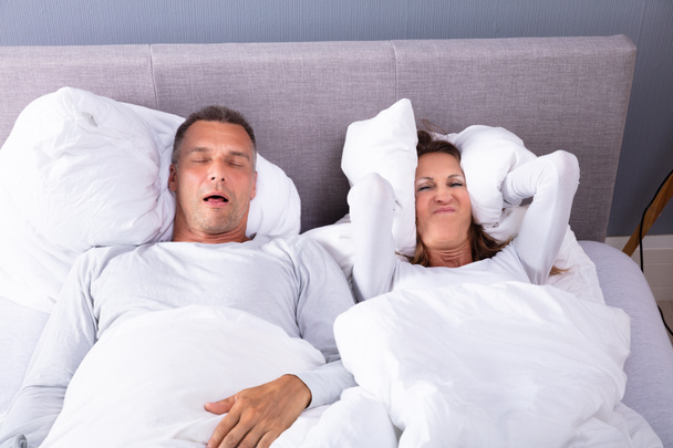 Femme mûre couvrant ses oreilles avec coussin tandis que l'homme ronfle au lit
 - Photo, image