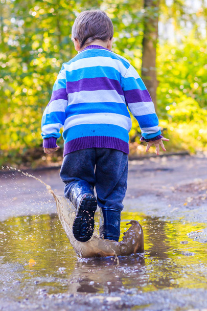 Ένα αγοράκι πηδάει σε μια λακκούβα. Ένα αγόρι με λαστιχένιες μπότες. Καλή παιδική ηλικία. Λιμνούλες μετά τη βροχή. Ζεστό καλοκαιρινό βράδυ. - Φωτογραφία, εικόνα