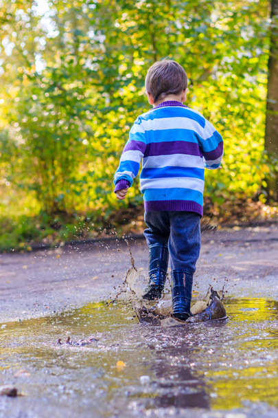 Ένα αγοράκι πηδάει σε μια λακκούβα. Ένα αγόρι με λαστιχένιες μπότες. Καλή παιδική ηλικία. Λιμνούλες μετά τη βροχή. Ζεστό καλοκαιρινό βράδυ. - Φωτογραφία, εικόνα