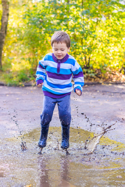 A little boy is jumping in a puddle. A boy in rubber boots. Happy childhood. Puddles after the rain. Warm summer evening. - Foto, Imagem