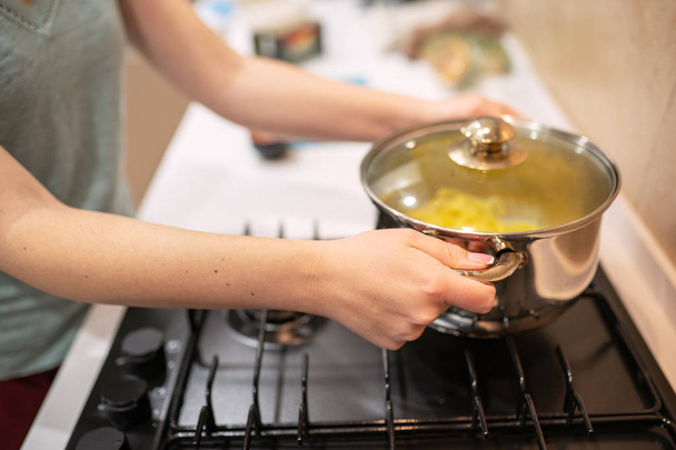 Beautiful young woman housewife prepairing dinner, hold in hands big steel saucepan, standing it on gas-stove. - Foto, Imagen