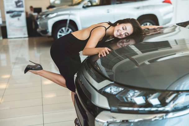 beautiful brunette woman in sexy black dress feeling happy hugging her brand new car in dealership center - Photo, Image