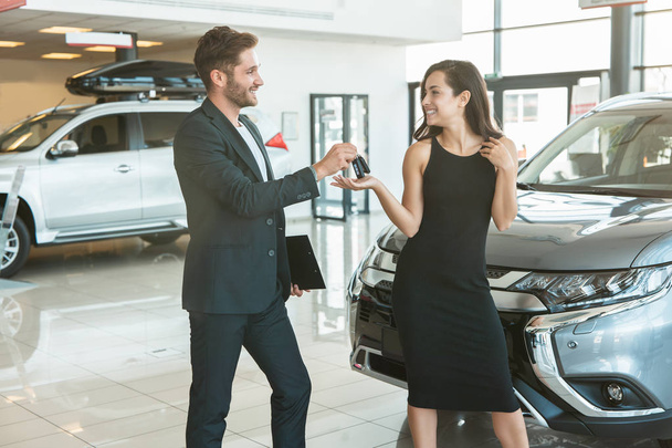 handsome man manager giving car keys to beautiful brunette woman client standing near brand new car after succesful deal in dealership center - Foto, afbeelding