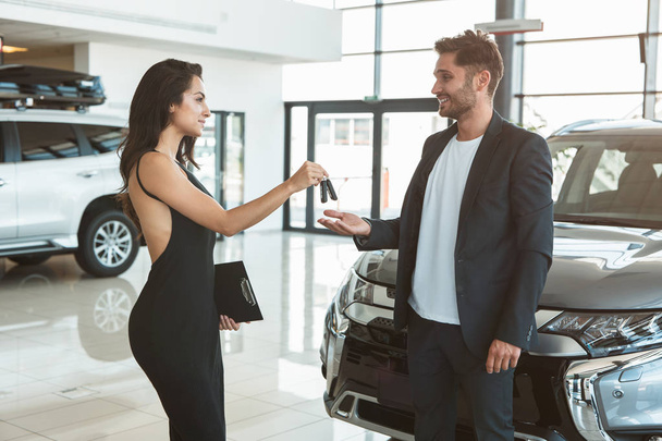 beautiful brunette woman manager giving car keys to handsome man client standing near brand new car after succesful deal in dealership center feeling happy - Foto, afbeelding