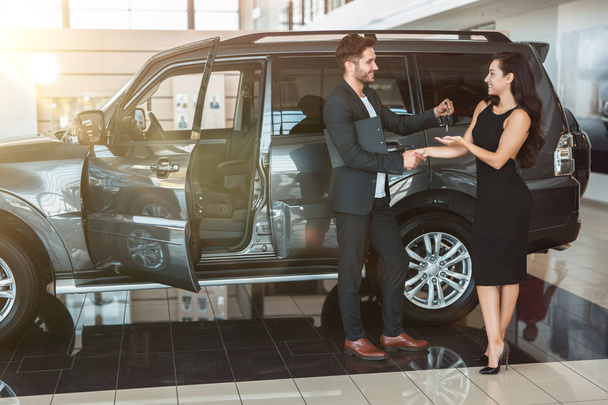 handsome man manager gives car keys to beautiful brunette woman client after succesful deal in dealership center standing near new SUV - Fotografie, Obrázek
