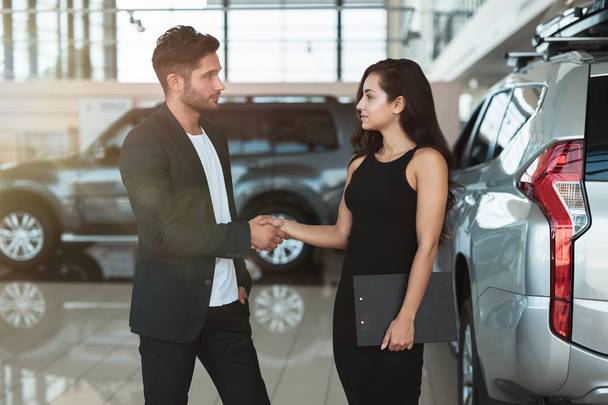 beautiful brunette woman manager shaking hands with handsome man client standing in dealership center after succesful deal - Photo, image