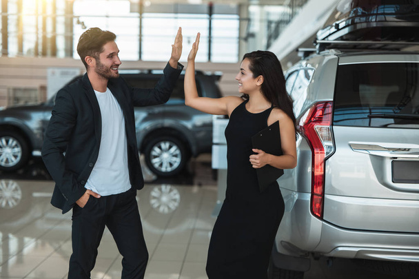 handsome man manager and beautiful brunette woman client standing near brand new SUV after succesful deal in dealership center - Foto, Imagen