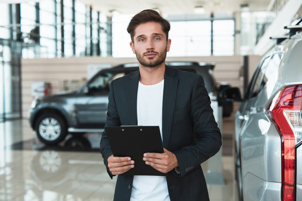 jonge knappe man Manager glimlachend houden brochure in zijn handen staande in dealer Center in de buurt van gloednieuwe SUV - Foto, afbeelding
