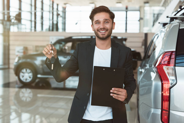 young handsome man manager smiling with brochure in his hand standing in dealership center near brand new SUV holding car keys - Photo, image