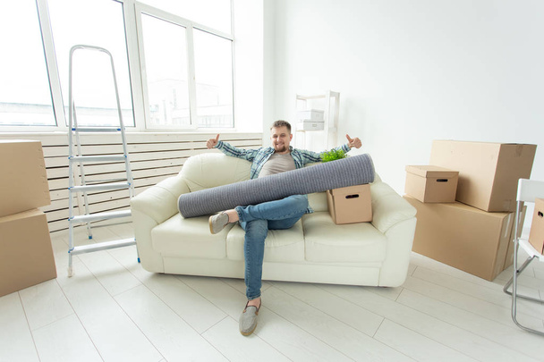 Позитивный молодой человек, сидящий на диване с вещами в новой гостиной после переезда. Концепция доступности жилья для молодежи
. - Фото, изображение