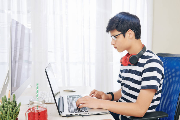 Серйозний хлопчик змішаної раси в окулярах з навушниками навколо шиї працює на ноутбуці в своїй кімнаті
 - Фото, зображення