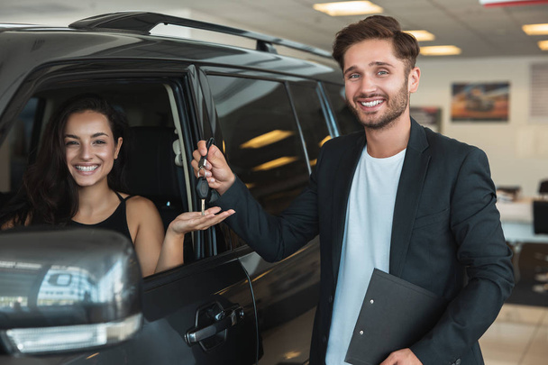 handsome man manager gives car keys to beautiful brunette woman client after succesful retail deal in dealership center standing near new SUV - Фото, зображення