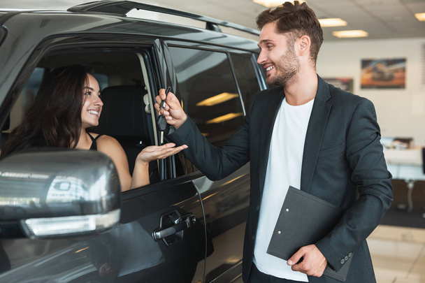 handsome man manager gives car keys to beautiful brunette woman client after succesful retail deal in dealership center standing near new purchased car - Foto, afbeelding