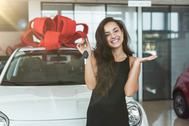 young beautiful woman feels happy holding keys from her brand new car standing in dealership center - Foto, Bild