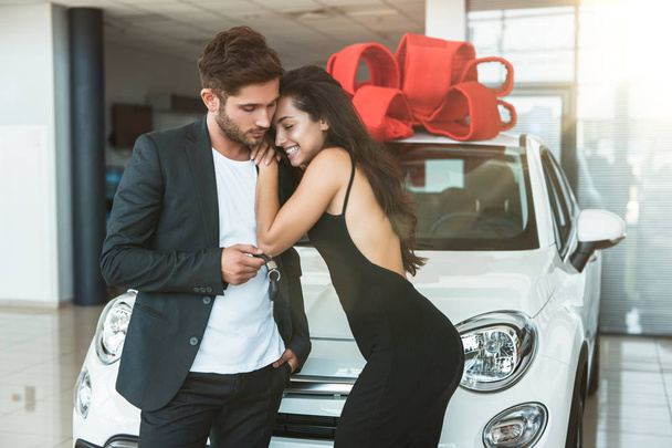 young beautiful woman looks grateful receiving brand new car for present from her boyfriend hugging him in dealership center - Fotó, kép