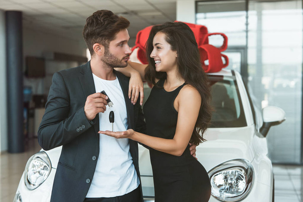 young handsome man gives keys from brand new car he presented to his beautiful woman standing in dealership center looking happy - Фото, зображення