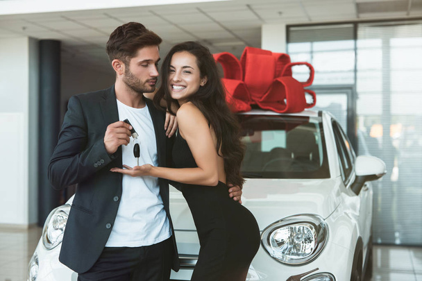 young handsome man gives keys from brand new car he presented to his beautiful wife standing in dealership center looking happy - Foto, afbeelding