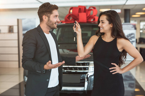young beautiful woman presents brand new car to her surprised handsome husband standing in dealership center - Valokuva, kuva