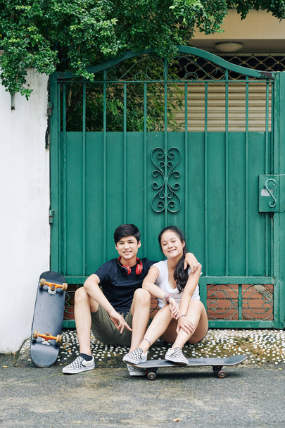 Улыбающиеся обнимающиеся подросток брат и сестра сидят перед воротами дома и отдыхают после скейтборда
 - Фото, изображение