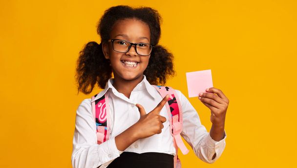 Μαύρη δημοτικού μαθητή κορίτσι δείχνοντας σε κενό χαρτί, κίτρινο φόντο - Φωτογραφία, εικόνα