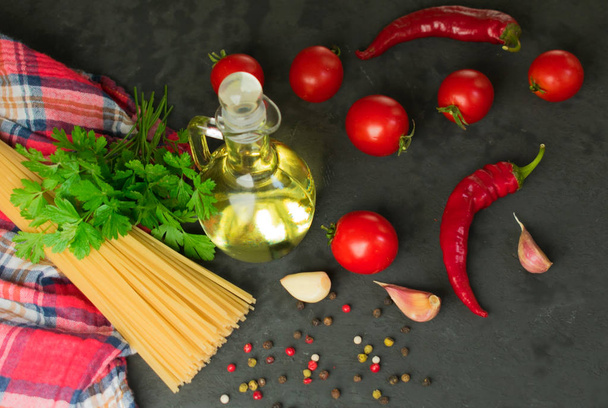 Ingredients for cooking spaghetti - raw spaghetti, cherry tomatoes, chili peppers, garlic, herbs, spices and olive oil on a black background. Culinary background. Selective focus. - Photo, image