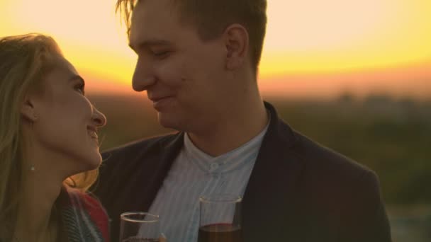 Man and young pretty woman sitting on couch, holding wine glasses and kissing on rooftop terrace at sunset. - Footage, Video
