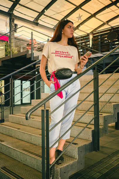 Πορτρέτο ενός κοριτσιού που ποζάρει κοντά σε ένα εμπορικό κέντρο, κρατώντας smartphone. Φόρεμα λευκό t-shirt, μπλε παντελόνι, μαύρη τσάντα μέσης και sneakers. - Φωτογραφία, εικόνα