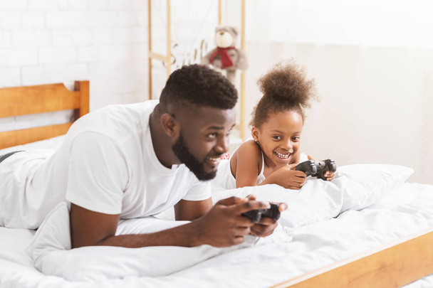 Afrikaanse man en zijn kleine meisje spelen op console met joysticks - Foto, afbeelding