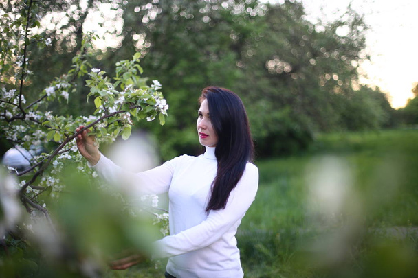Молода усміхнена жінка з довгим чорним волоссям у білій черепашці весна квітуча бузкова і яблуня
 - Фото, зображення