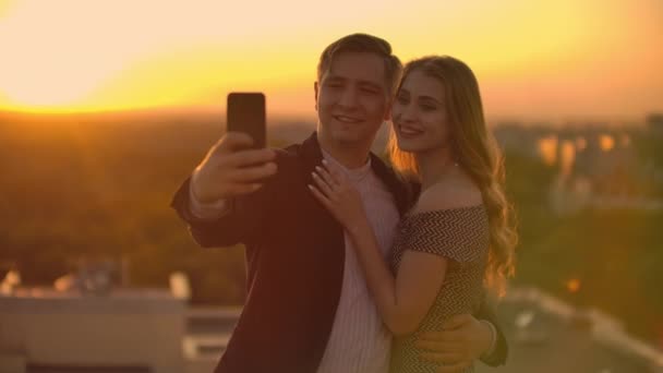 Staande op het dak bij zonsondergang een getrouwd stel een man en een vrouw knuffel en neem een selfie op de telefoon. Maak foto's van jezelf staande op het dak en knuffel. Jonge mensen verliefd. - Video