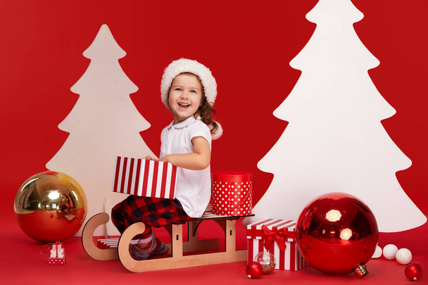 Merry Christmas and Happy Holidays! Cute little child girl is decorating the Christmas tree indoors. Isolated on red background. A child sits on a sleigh and opening gift boxes - Zdjęcie, obraz