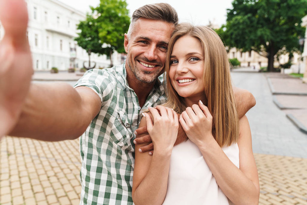 Image of charming young couple smiling and taking selfie photo together while walking through city street - Foto, immagini
