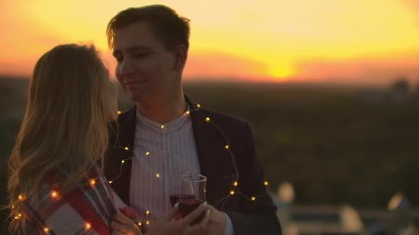 Man and young pretty woman sitting on couch, holding wine glasses and kissing on rooftop terrace at sunset. - Footage, Video